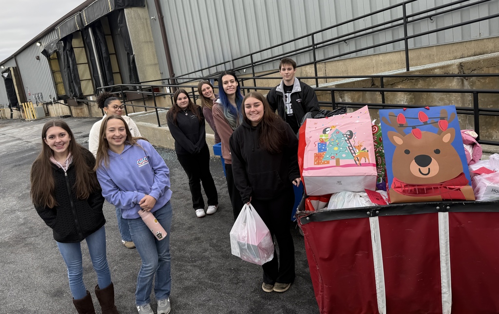 A group of nine students stand outside a warehouse beside a large cart filled with holiday-themed gift bags. They are smiling for the photo with the building and loading dock ramps visible behind them.
