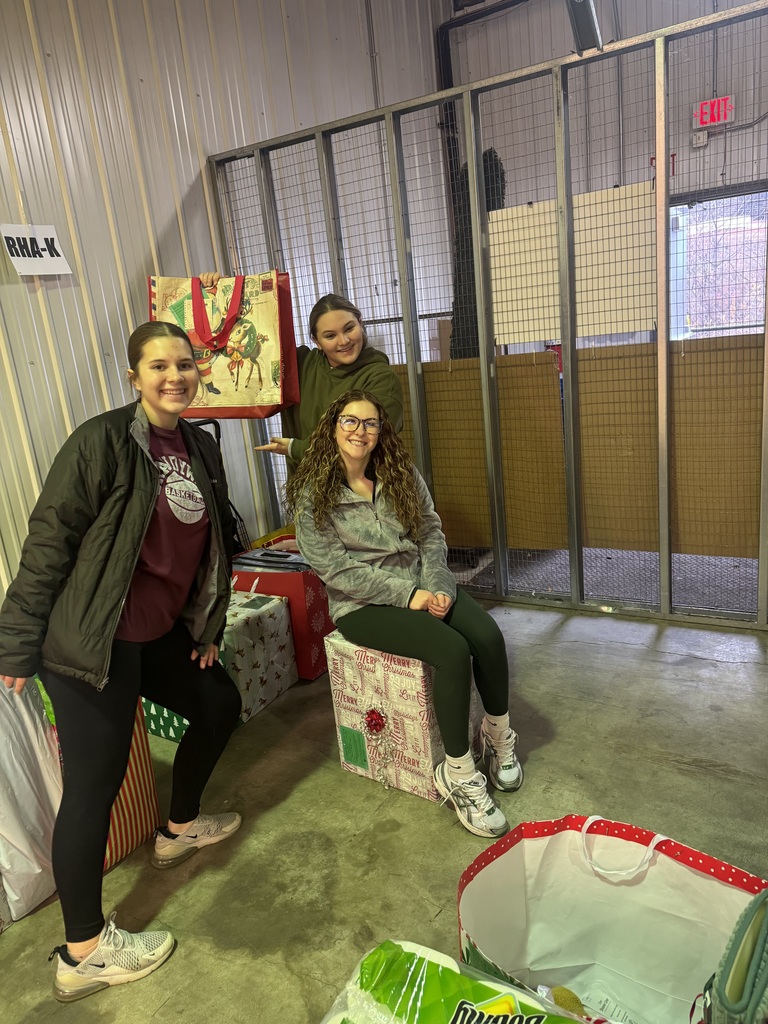 Three students are inside a warehouse sorting holiday donations. Two students stand while one sits on a wrapped box, all smiling. Large decorated gift bags and section signs on wire partitions are visible behind them.