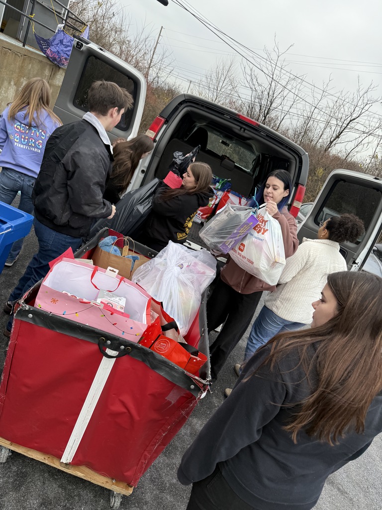 Multiple students unload holiday donation bags from a vehicle outside a warehouse loading dock. They are carrying gift bags and packages while working together around a large red cart full of donations.