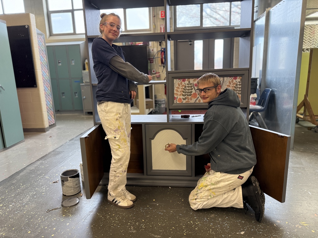 Two students pose beside a refinished cabinet with gray paint and patterned paper accents. One student stands while the other kneels and installs a drawer pull. Both wear safety glasses and paint-splattered pants in a studio environment.