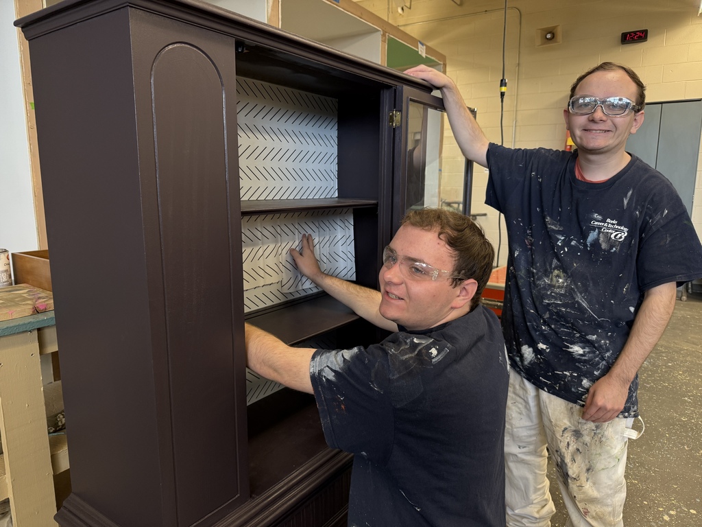 Two Residential Painting & Interior Design students stand beside a dark-painted cabinet. One student kneels inside the cabinet applying patterned paper to the back panel while the other stands smiling with safety glasses on. Both wear paint-splattered clothing in a workshop setting.