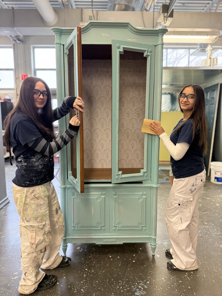 Two students stand beside a tall teal cabinet with decorative trim. One holds the door open while the other sands the surface. Both wear safety glasses and paint-marked work clothes in a classroom workshop.
