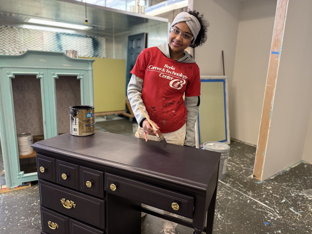 A student wearing safety glasses, a headband, and a red Berks Career & Technology Center shirt paints a dark-colored desk in a workshop. The desk has multiple drawers with gold knobs, and paint supplies are visible in the background.