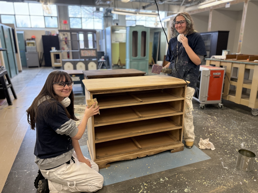 Two students sand a wooden dresser, smiling toward the camera. One kneels while the other stands with sanding tools in hand. The workshop background includes refinished furniture pieces and paint supplies.