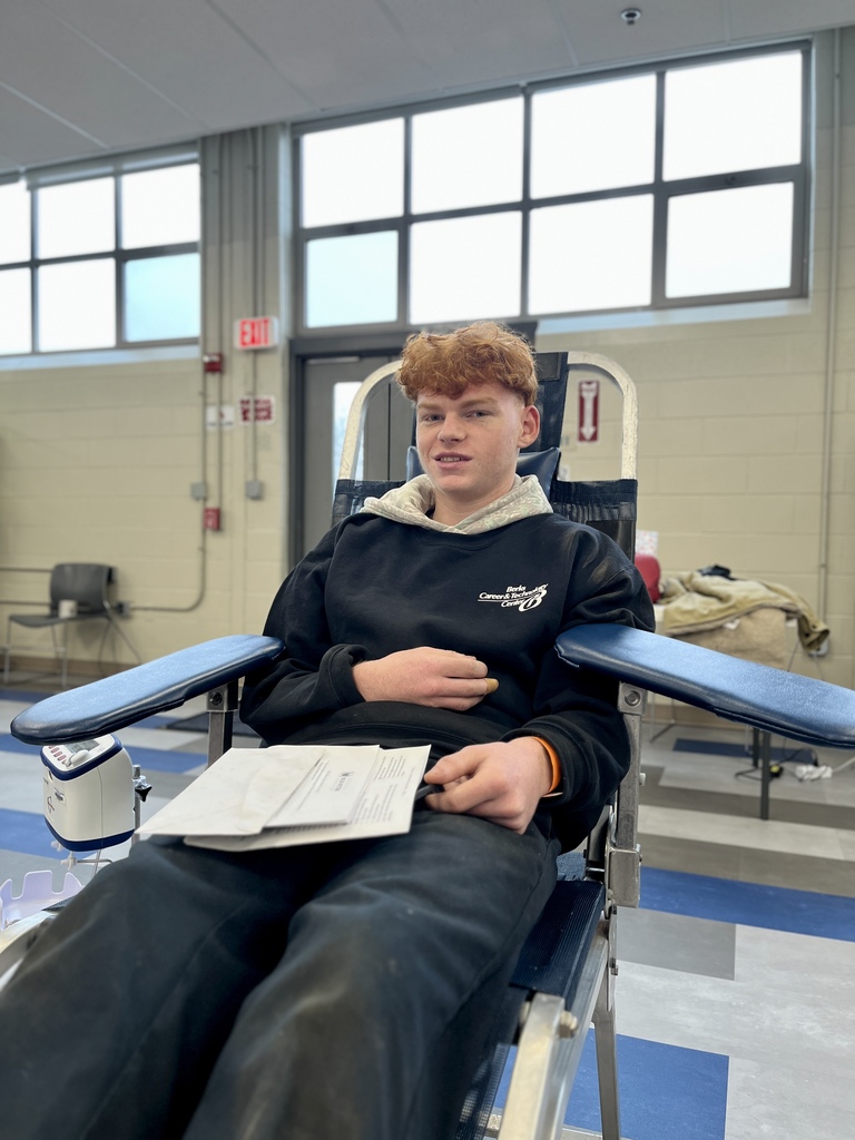 A student with red hair reclines in a donation chair, smiling slightly while holding paperwork. The classroom background includes windows, chairs, and light holiday décor.