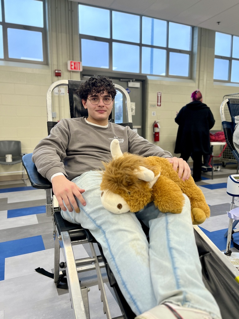A student reclines in a donation chair smiling with a plush stuffed llama across his lap. Classroom windows and lab equipment are seen in the background.