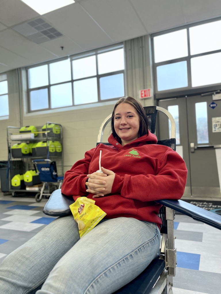 A student in a red hoodie reclines in a donation chair, smiling with a drink and snack resting on her lap. Large classroom windows and equipment storage racks are visible in the background.