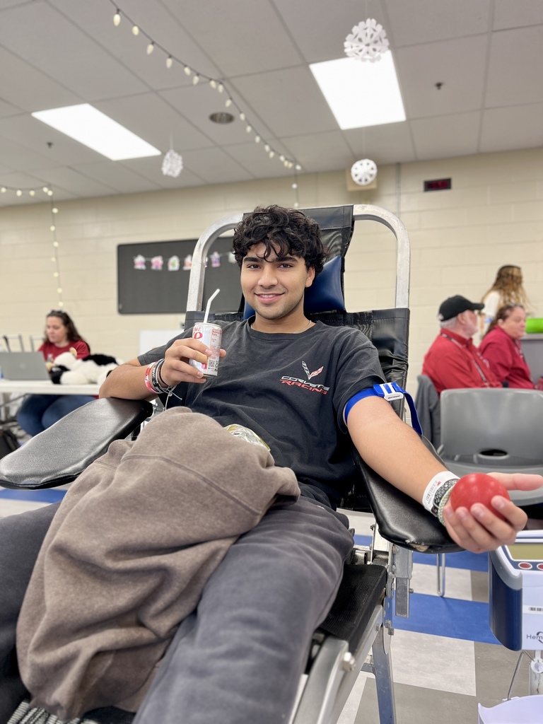 A student reclines in a donation chair, smiling while holding a juice box and squeezing a red stress ball. Other donors and staff are visible in the softly blurred background under hanging holiday decorations.