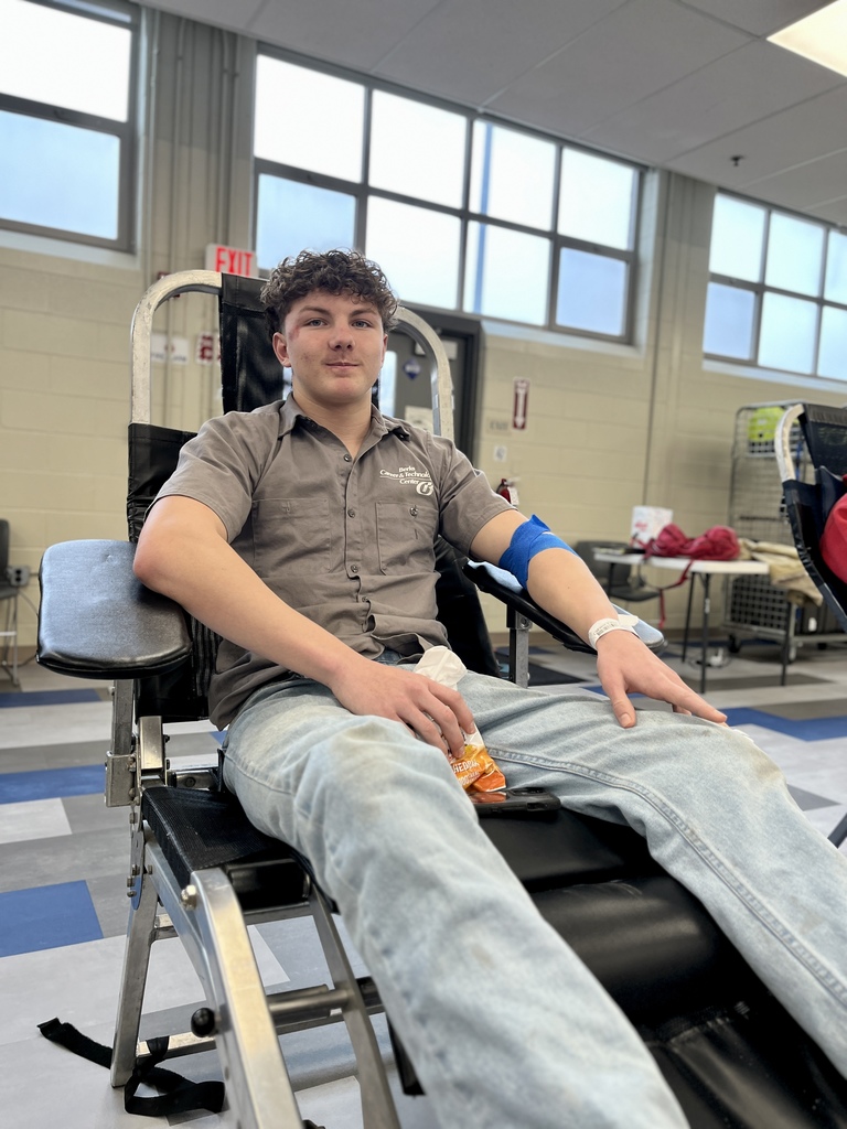 A student wearing a BCTC shirt sits in a donation chair with a blue bandage on his arm, holding a snack. Bright classroom windows and lab equipment appear in the background.
