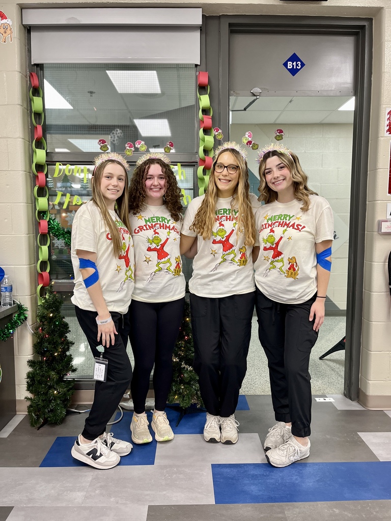 Four Health Occupations students pose together wearing matching “Merry Grinchmas” shirts and festive headbands. They stand in front of decorated classroom doors with holiday garland and miniature Christmas trees.