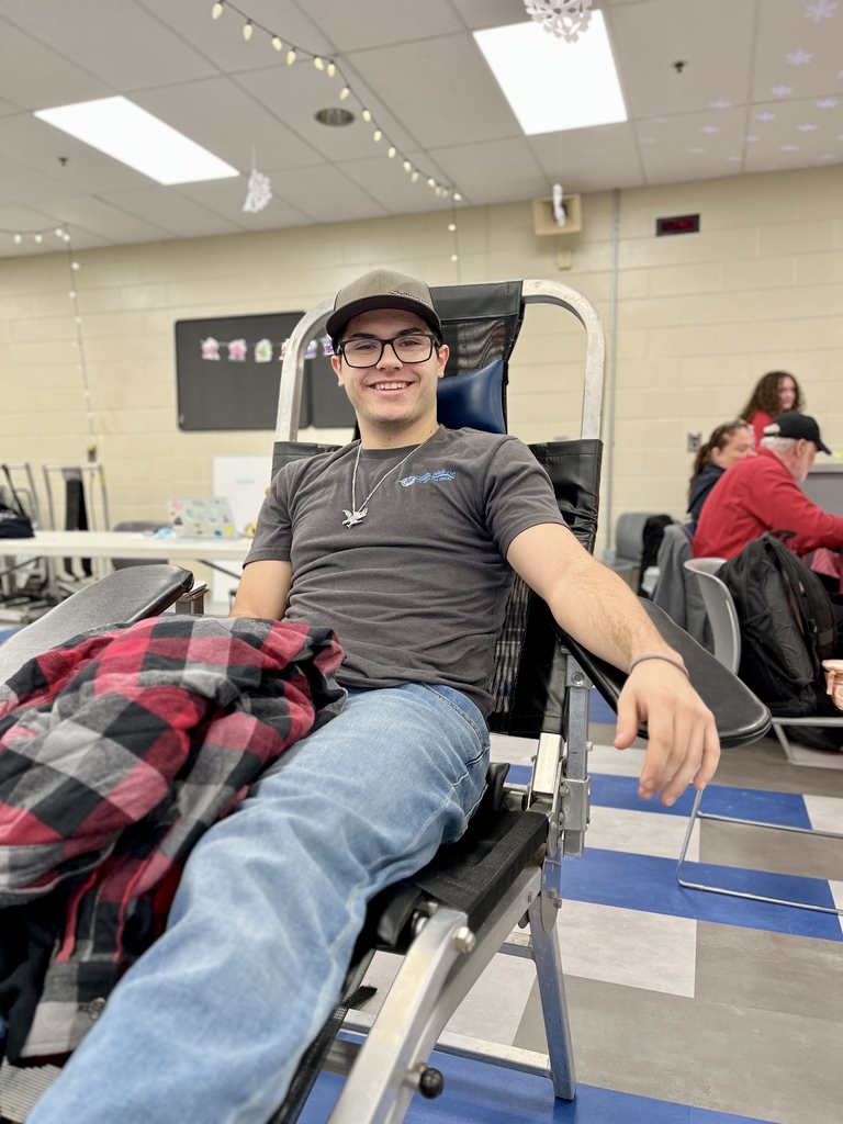 A student wearing glasses reclines in a donation chair smiling, with a flannel jacket draped over their lap. Other students and donors are visible in the background.
