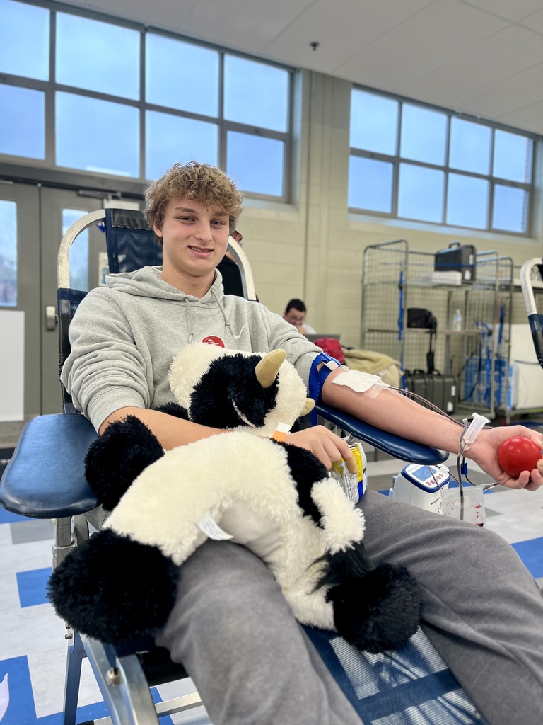 A student reclines in a donation chair with an IV in his arm, holding a stress ball while hugging a large stuffed cow. Classroom windows and carts are visible in the background.