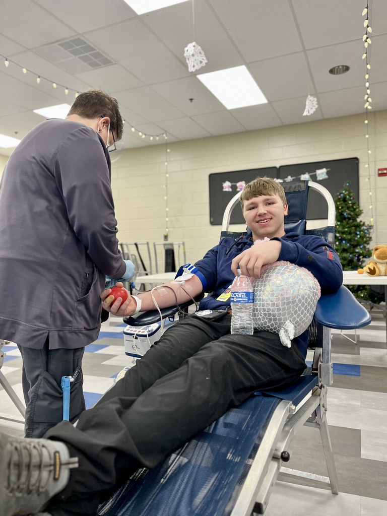 A student reclines in a donation chair with an IV in his arm while a healthcare worker assists beside him. He holds a water bottle and a wrapped ham on his lap. Holiday lights and a decorated tree are visible in the background.