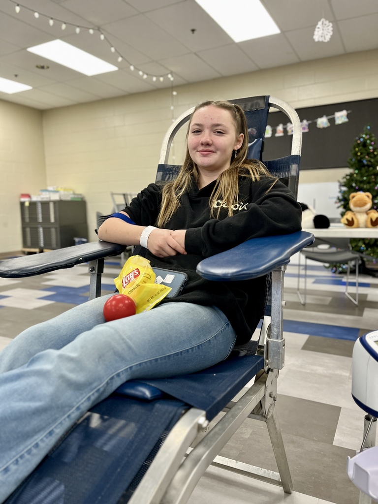 A student sits confidently in a reclining donation chair after giving blood, holding snacks including a bag of chips and an apple stress ball. The setting is a decorated classroom with holiday lights and a Christmas tree blurred in the background.