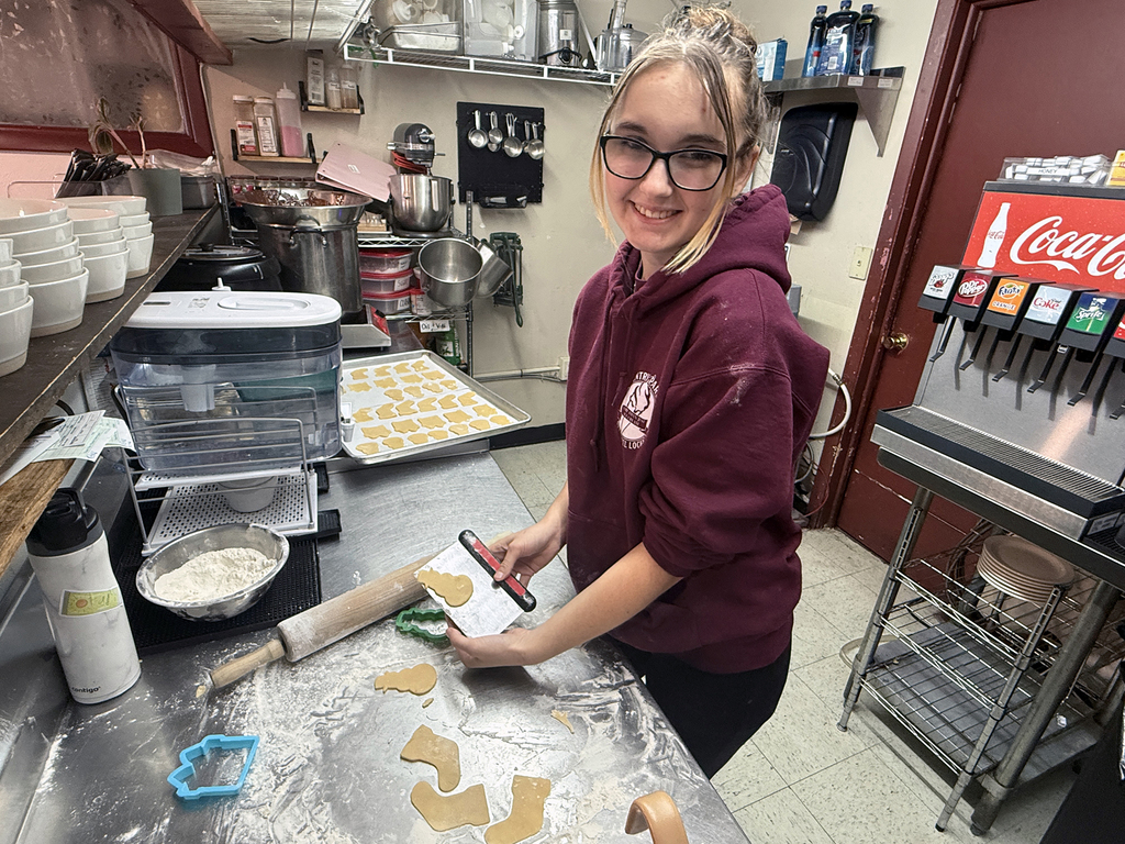 A high school student wearing glasses and a maroon Coventry Parlor at Laurel Locks hoodie smiles while cutting cookie dough in a commercial kitchen. She stands at a stainless-steel counter dusted with flour, using a rolling pin, scraper, and cookie cutters to shape the dough. A tray of unbaked cookies sits behind her, and shelves with kitchen tools, bowls, and equipment are visible in the background.