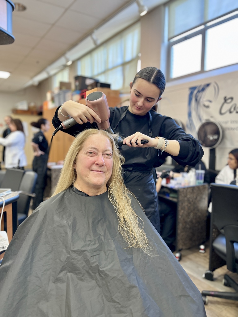 A cosmetology student blow-dries a client’s hair while the client sits in a salon chair wearing a black cape, with other students working in the background.