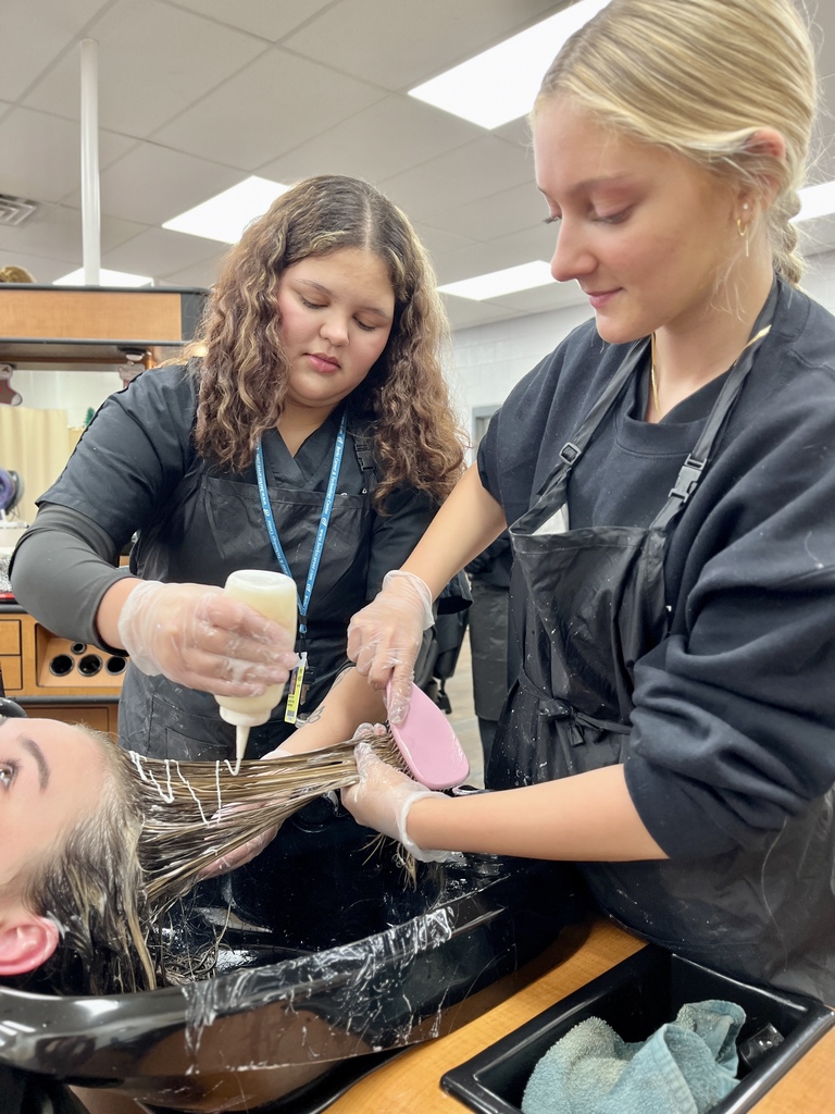 Two cosmetology students apply hair color to a client’s hair at a shampoo bowl, carefully sectioning and saturating strands in a classroom salon.