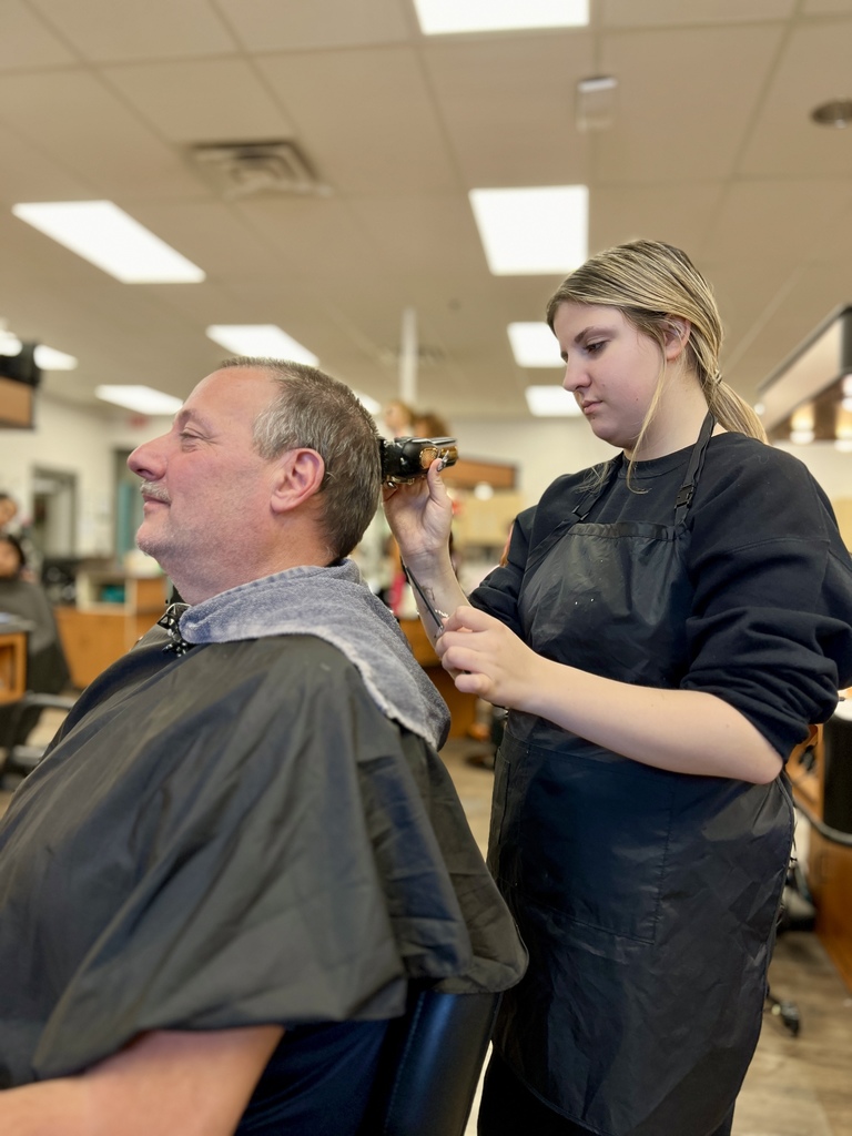 A cosmetology student trims a client’s hair using clippers at the back of the head in a classroom salon setting.