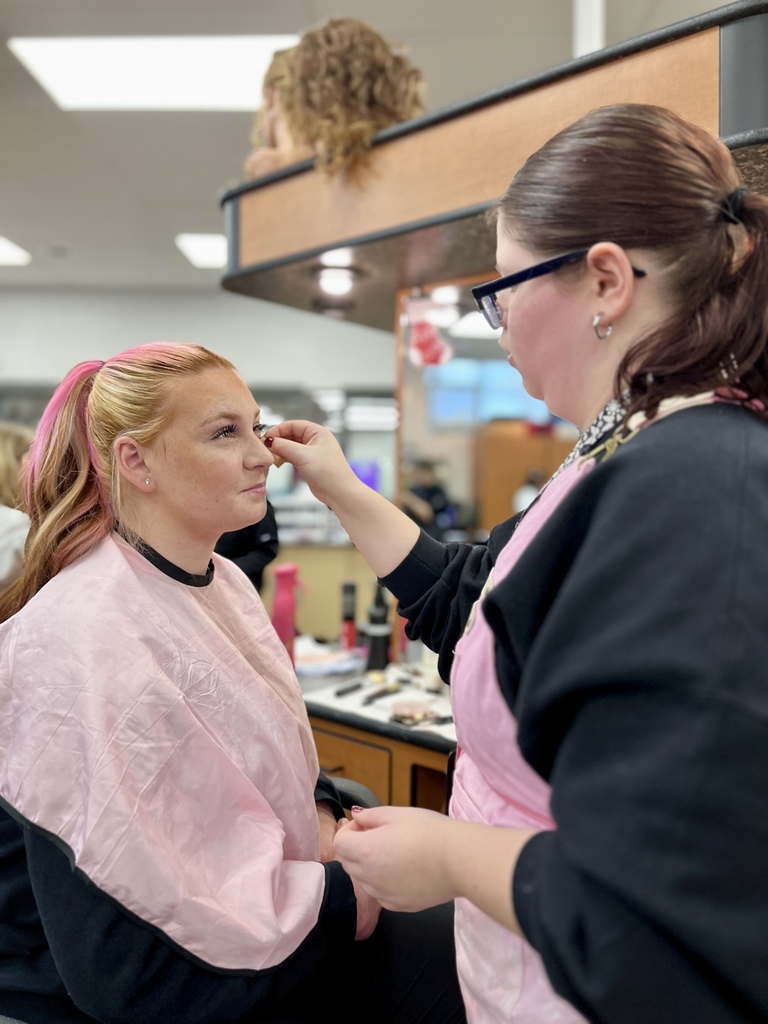 A cosmetology student applies eye makeup to a seated client wearing a pink salon cape, with styling stations visible behind them.