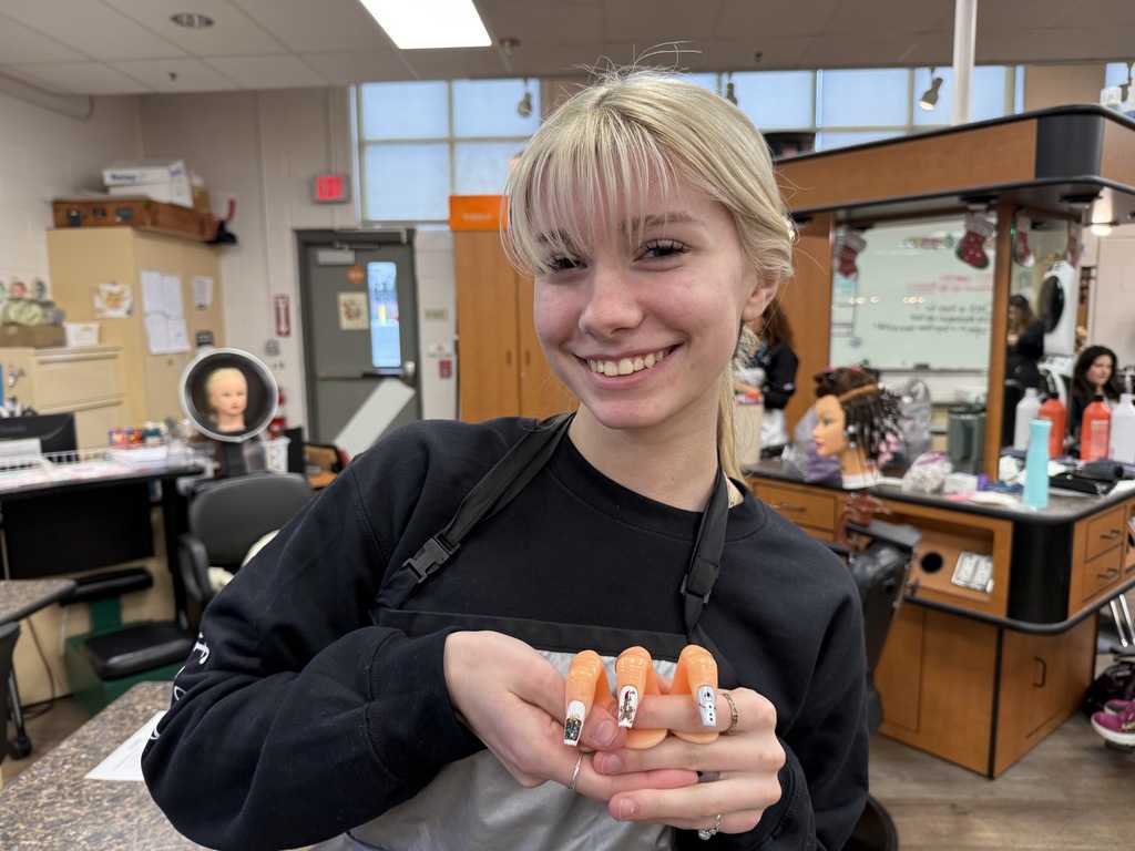 A cosmetology student smiles while holding festive nail art designs displayed on small practice nails in a classroom salon.