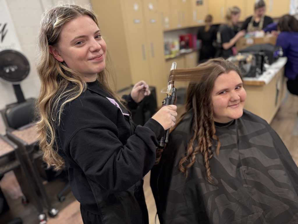 A cosmetology student uses a curling iron to style a client’s hair, creating defined curls while both face the camera in a salon classroom.