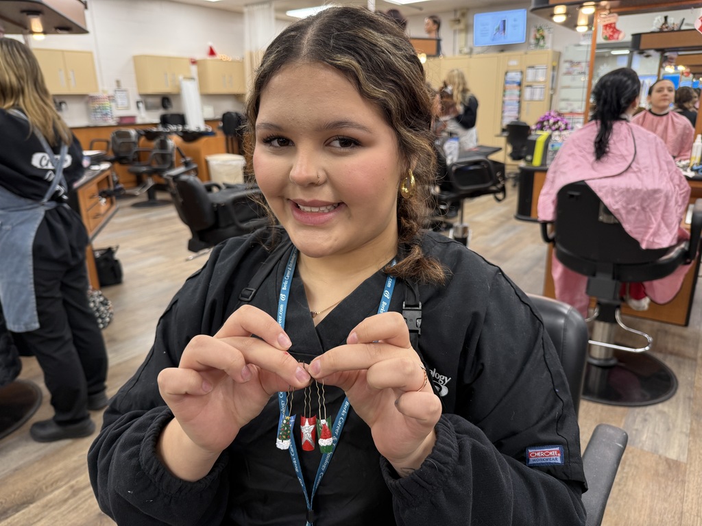 A cosmetology student smiles while holding festive nail art designs displayed on small practice nails in a classroom salon.