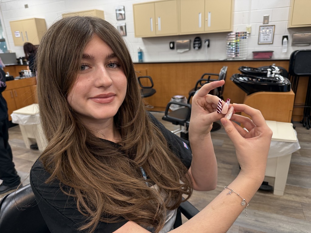 A cosmetology student smiles while holding festive nail art designs displayed on small practice nails in a classroom salon.