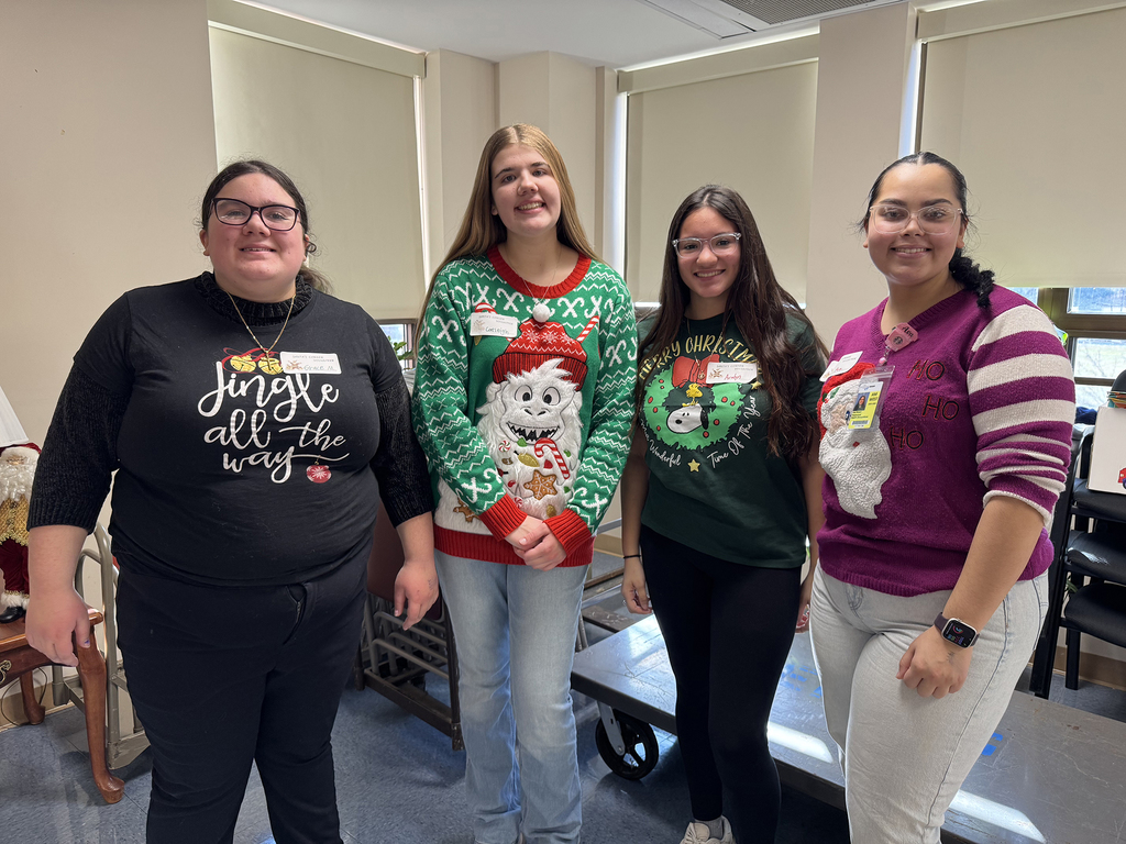 Group photo of Health Occupations students and staff seated and standing around a decorated chair, posing under a colorful balloon arch in a common area.