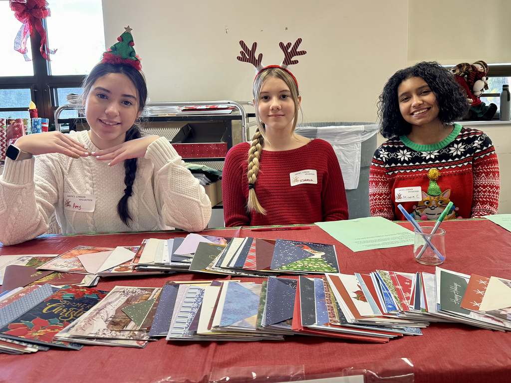 Three Health Occupations students wearing holiday headbands sit behind a table covered with stacks of holiday greeting cards and envelopes, smiling at the camera.