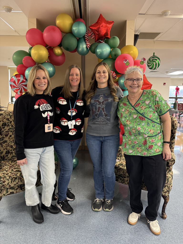 Two Health Occupations students sit behind a table covered with holiday cards and envelopes, smiling at the camera in a room with bookshelves in the background.