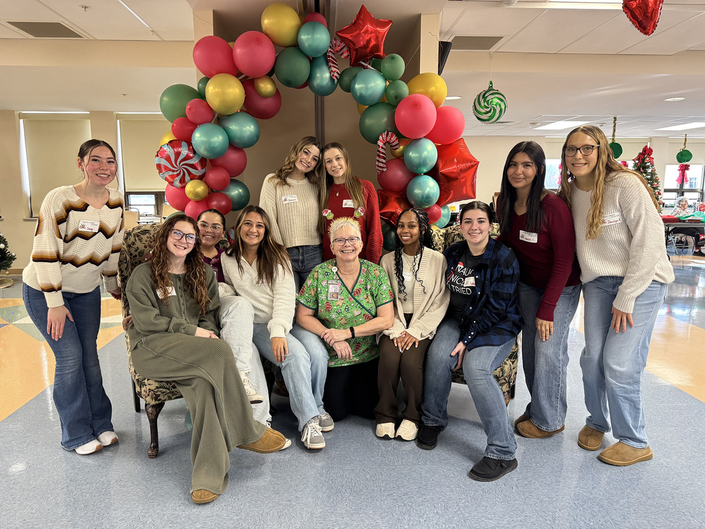 Four staff members stand side by side beneath a colorful balloon arch, smiling and wearing holiday-themed clothing.