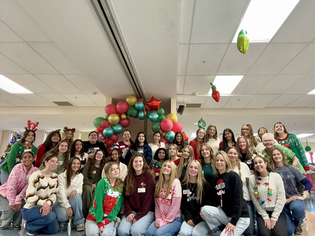 Group photo of Health Occupations students and instructors wearing festive holiday sweaters, posing together in a decorated common area with a colorful balloon arch and holiday decorations.
