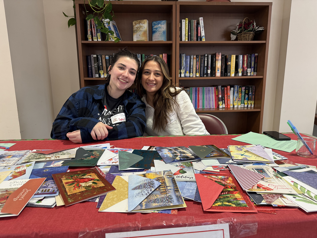 Two Health Occupations students stand beside a round table displaying small holiday gift items and figurines, smiling in a hallway decorated for the holidays.