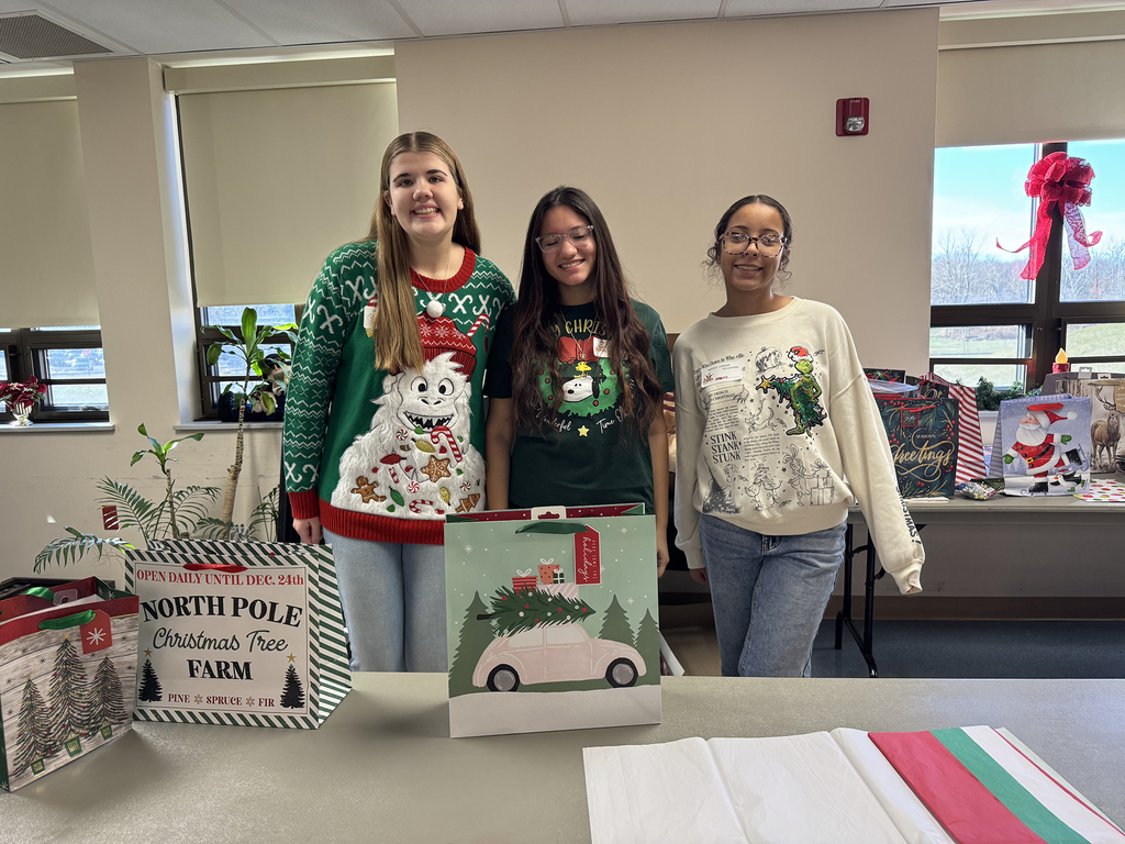 Three Health Occupations students stand behind a table with holiday gift bags, smiling at the camera in a festively decorated room.