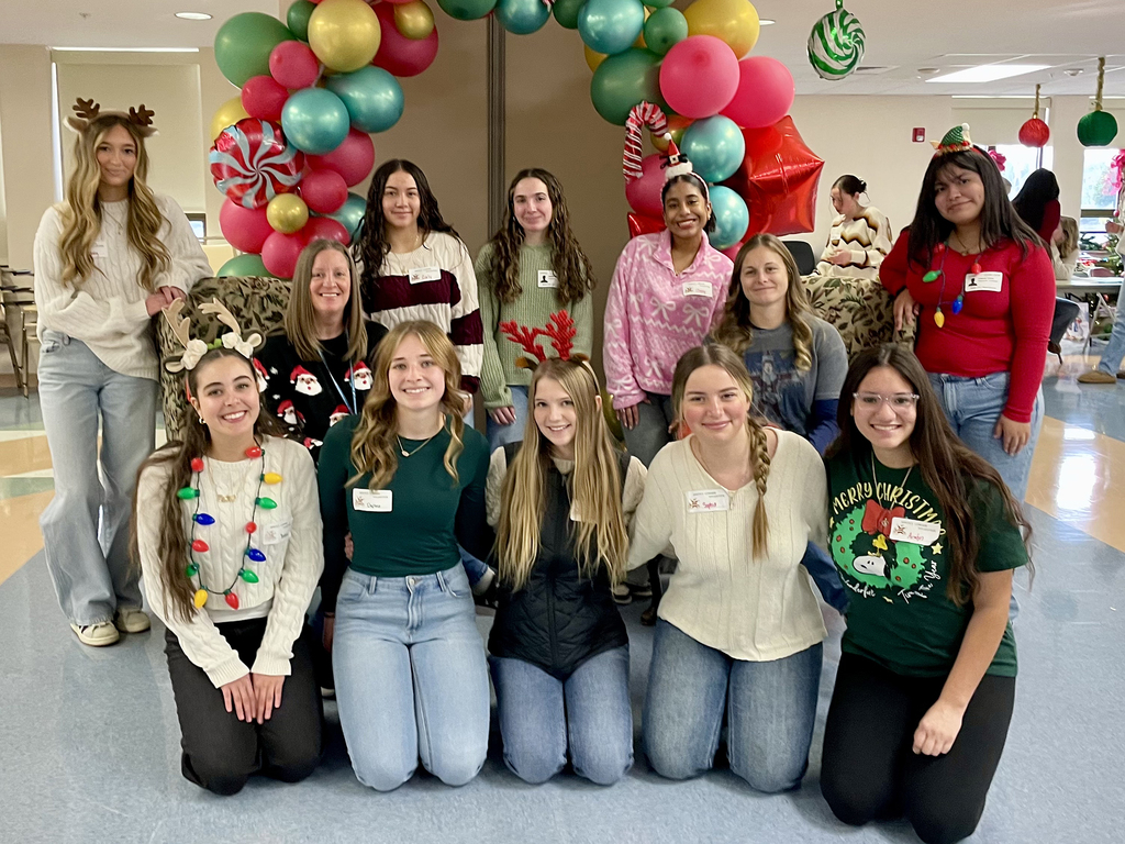 Large group of Health Occupations students and a staff member kneel and stand together, smiling in a festively decorated common space.