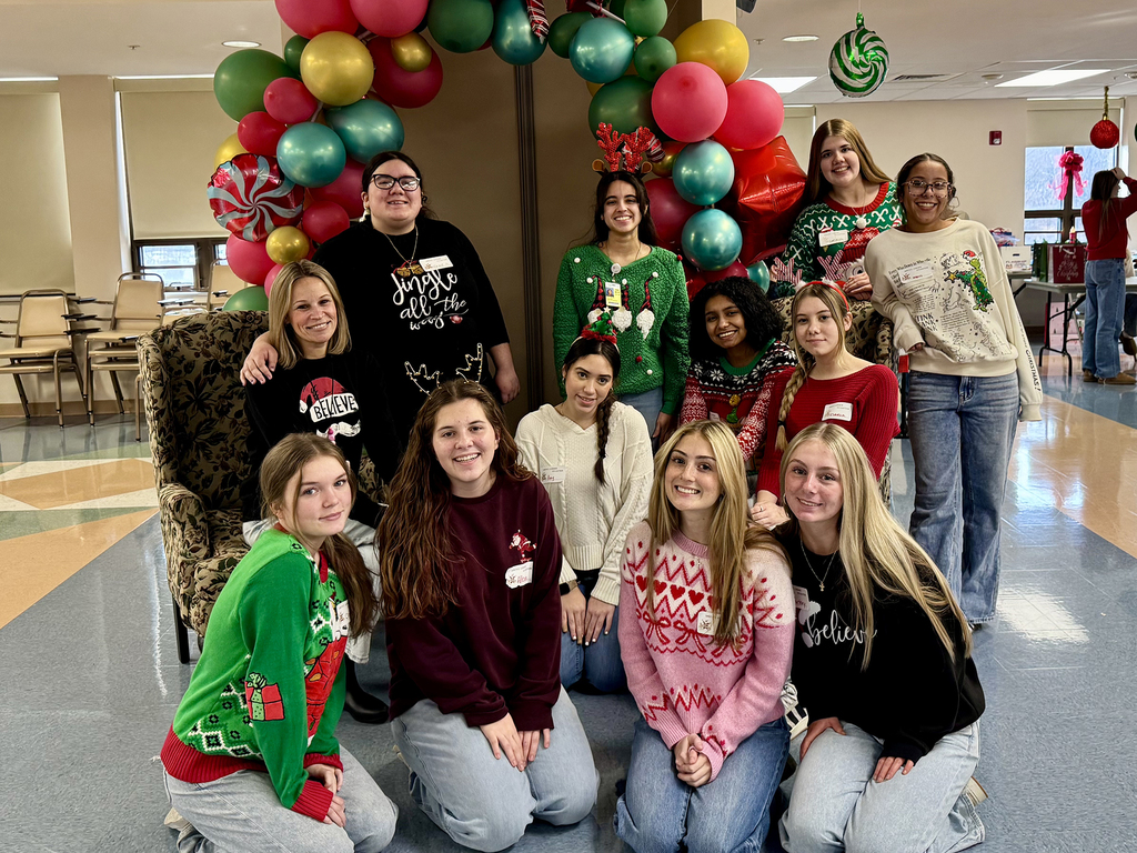 Large group of Health Occupations students and a staff member pose together indoors, wearing holiday attire and smiling in front of festive decorations.