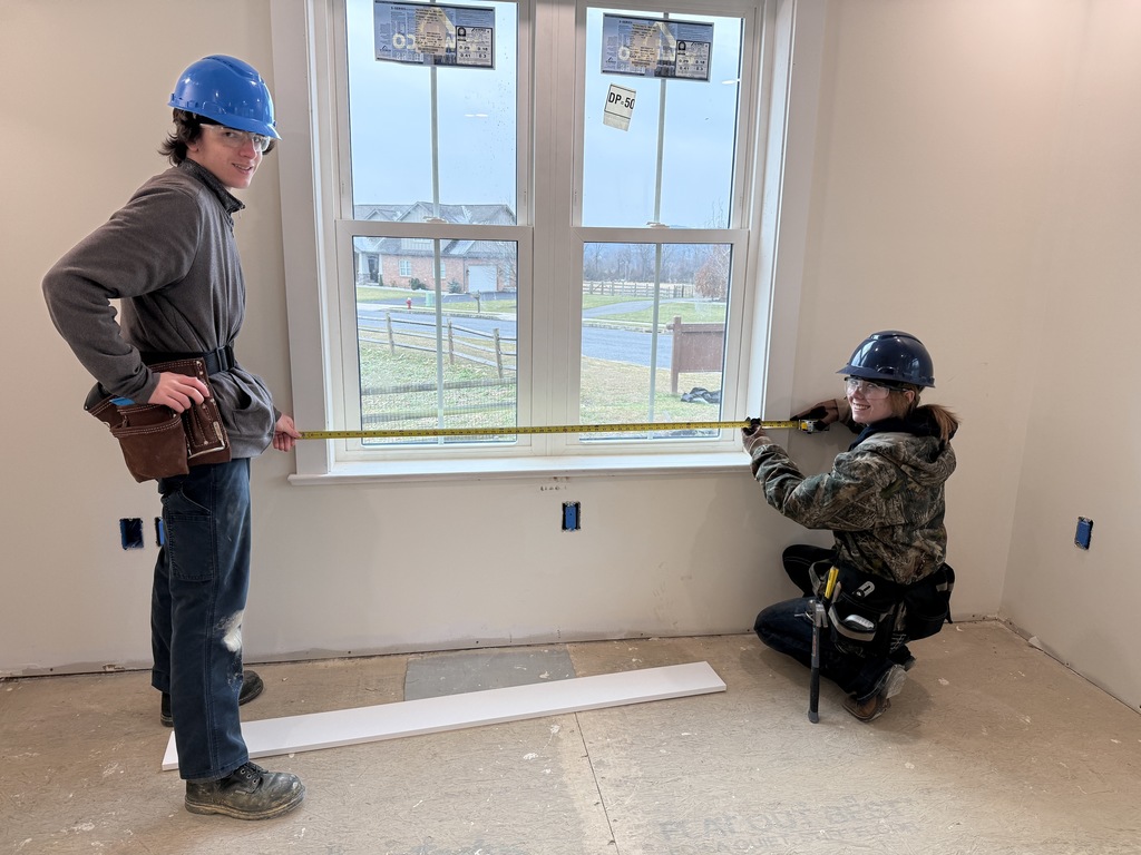 Carpentry students wearing hard hats measure the width of a window opening inside the student-built house, preparing for interior trim installation.