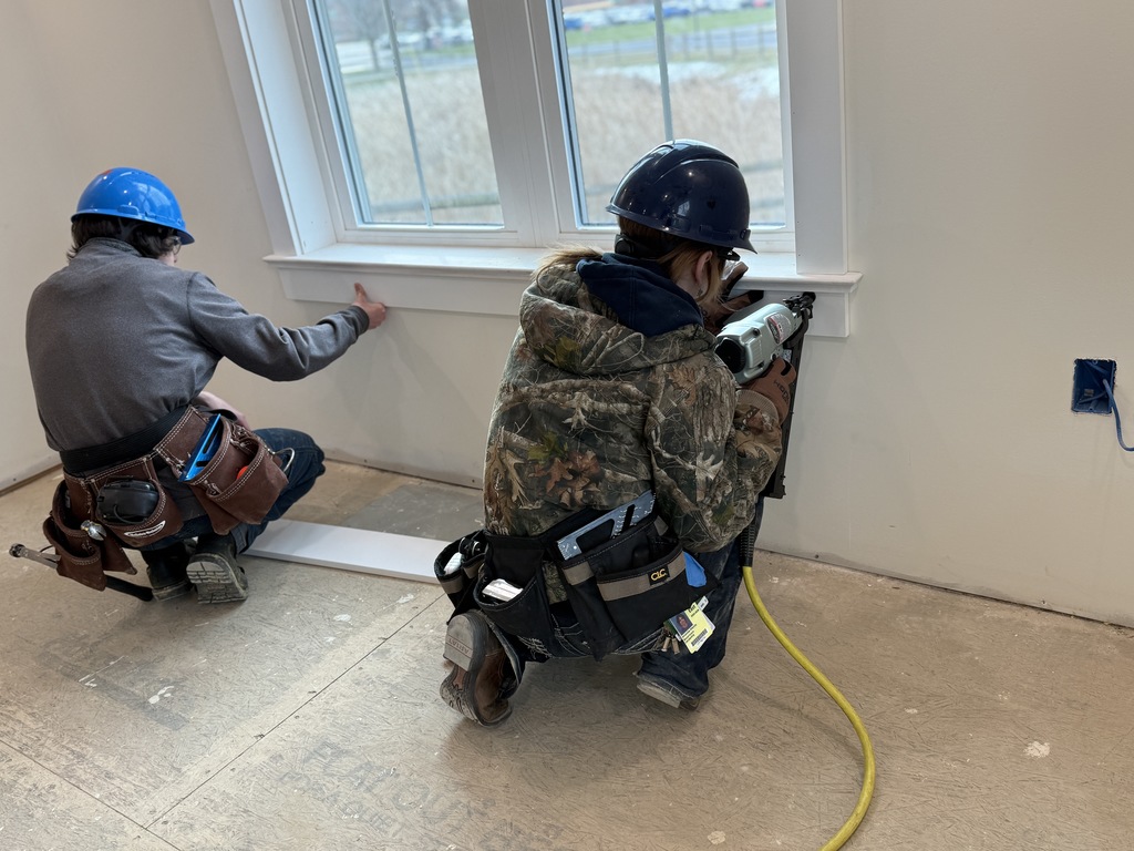 Two carpentry students kneel beneath a window while installing interior trim, using a nail gun and hand tools to secure the piece in place.