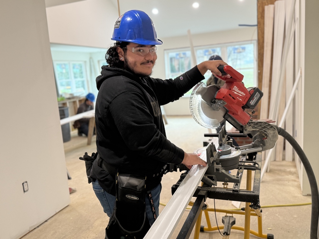A carpentry student wearing safety glasses and a hard hat positions interior trim on a miter saw, preparing to make a precise cut.