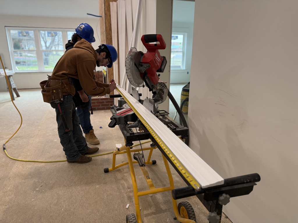 Two carpentry students measure and mark a long piece of interior trim positioned on a miter saw stand inside the student-built house.