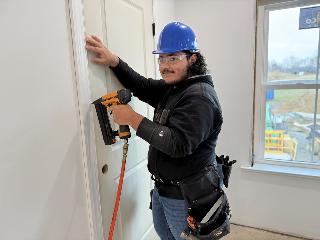 A carpentry student wearing a hard hat uses a nail gun to secure trim around an interior door frame inside the student-built house.