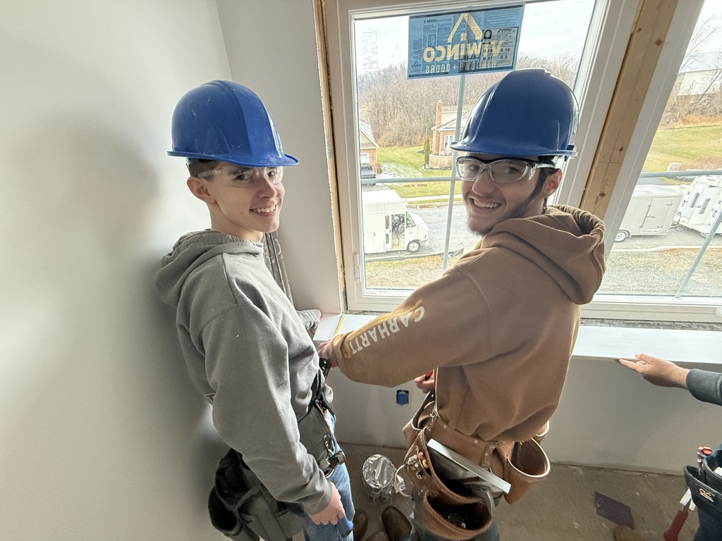 Two carpentry students smile toward the camera while positioning interior trim beneath a window during installation.