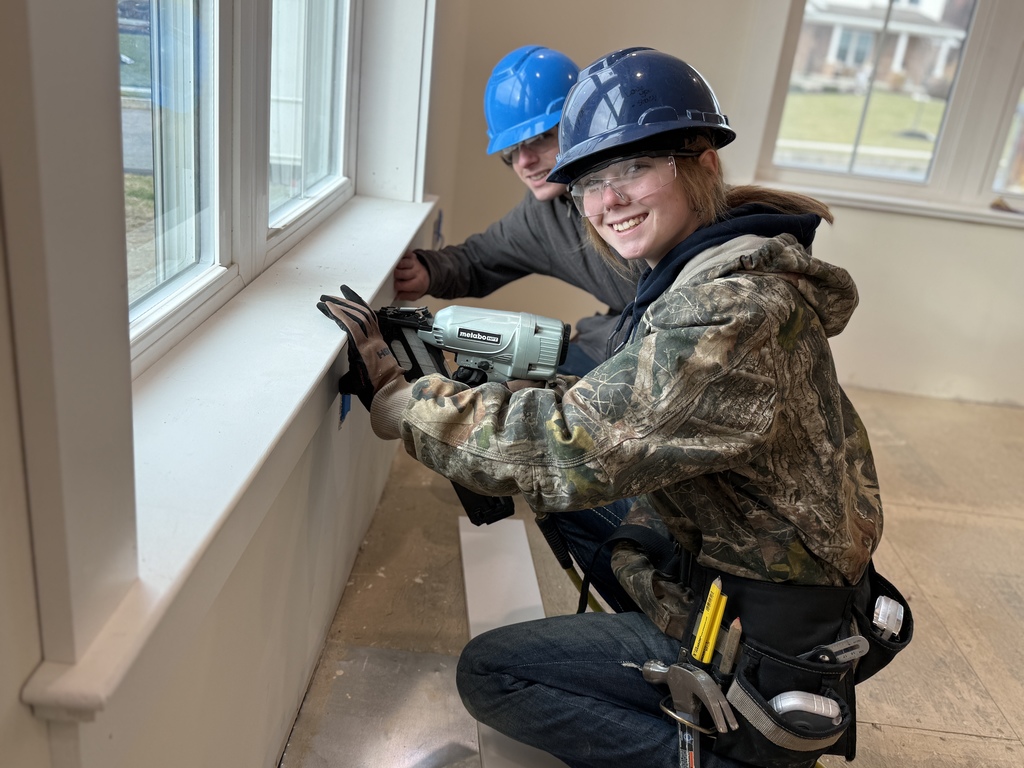 Two carpentry students kneel beneath a window while installing interior trim, using a nail gun and hand tools to secure the piece in place.