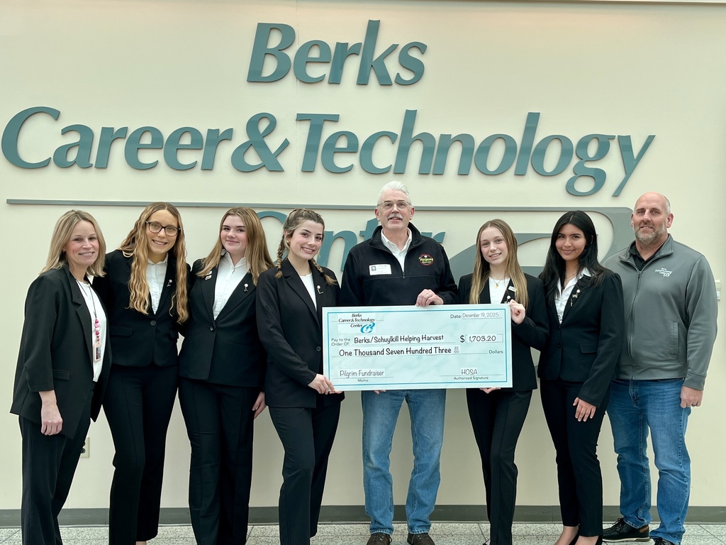 HOSA students from Berks Career & Technology Center stand with a Helping Harvest representative and staff members in front of a “Berks Career & Technology Center” wall sign, presenting an oversized check for $1,703.20 raised through their Pilgrim fundraiser.