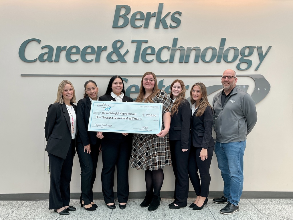 Group of Berks Career & Technology Center HOSA students and staff standing indoors in front of a wall sign reading “Berks Career & Technology Center,” holding an oversized donation check for $1,703.20 made out to Helping Harvest as part of a Pilgrim fundraiser.