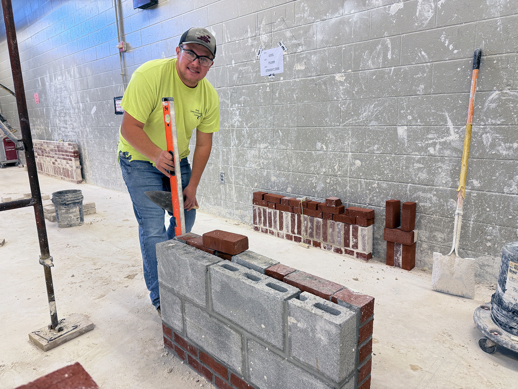 A student in a construction training lab uses a level to check the alignment of a partially built masonry wall made of concrete blocks and bricks, with tools and stacked bricks visible in the background.