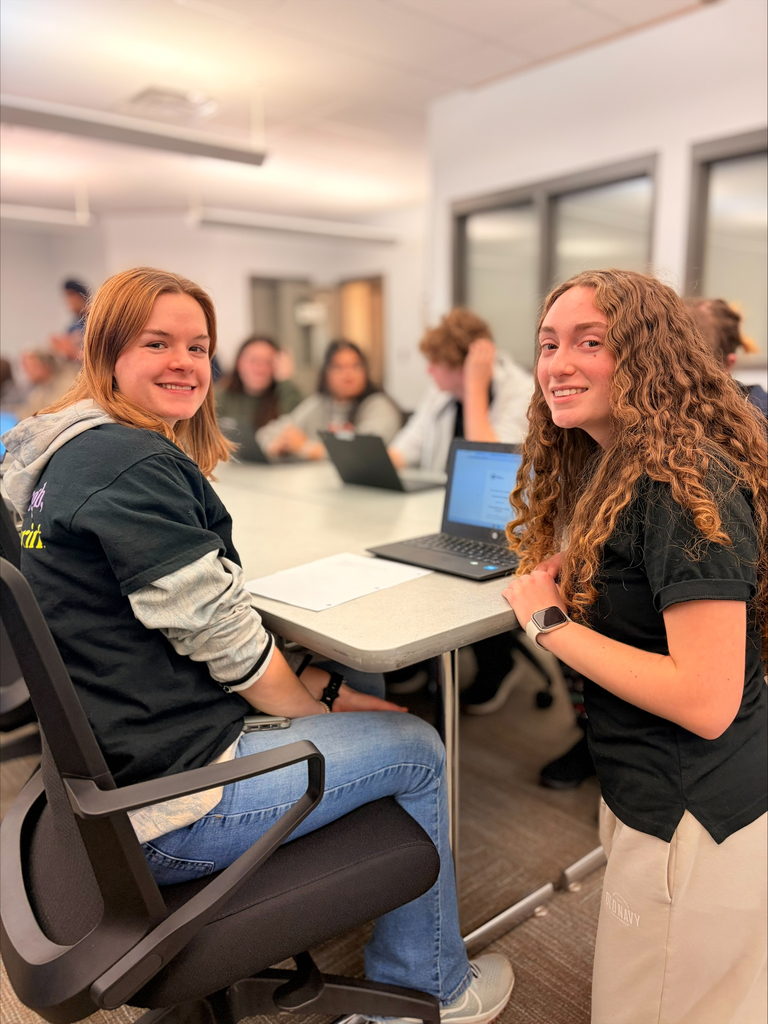 Two Business Management & Entrepreneurship students smile at the camera while sitting and kneeling beside a classroom table with laptops. Other students are visible working in the background during the Virtual Enterprises trade show activity.