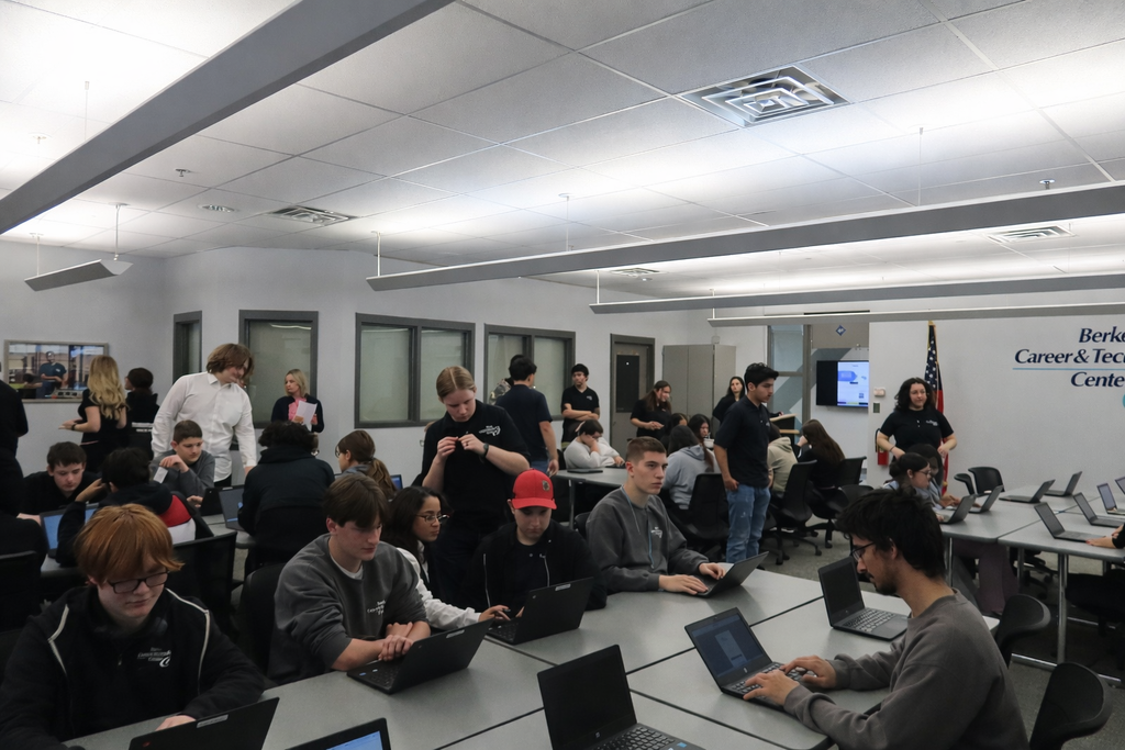 A large classroom at Berks Career & Technology Center filled with students sitting at tables working on laptops. Several Business Management & Entrepreneurship students stand and assist their peers around the room. The setting is bright and modern with gray tables, office chairs, and BCTC signage on the wall.