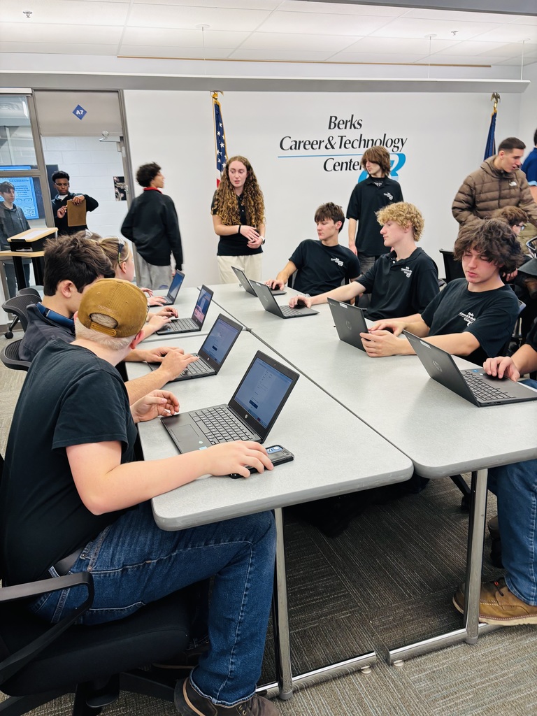 Students sit in rows at long tables working on laptops inside a BCTC classroom, while Business Management & Entrepreneurship students stand nearby to assist. The BCTC logo and American flag are visible on the wall in the background.