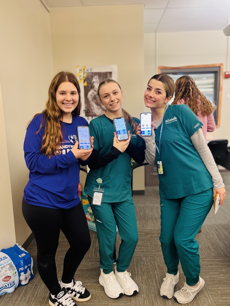 Three female students stand side-by-side smiling and holding up their smartphones toward the camera, each displaying an online shopping screen. Two students are wearing green medical scrubs and one is wearing a blue long-sleeve shirt. They appear to be participating as customers in the Virtual Enterprises trade show.